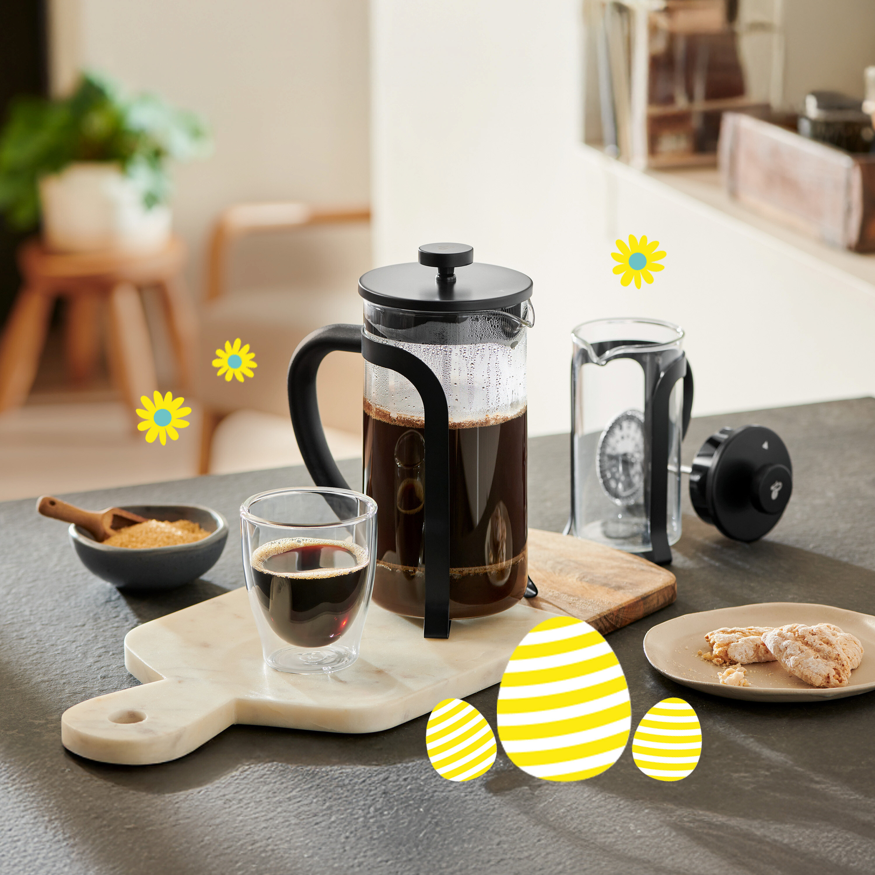 French press vedle šálku kávy, misky s cukrem a sušenek na stole se žlutými velikonočními vajíčky a sedmikráskami.