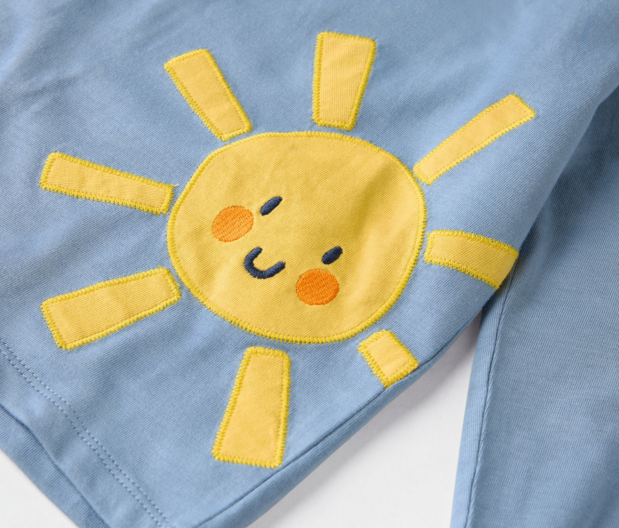 Detail světle modrého oděvu s našitým žlutým sluncem.