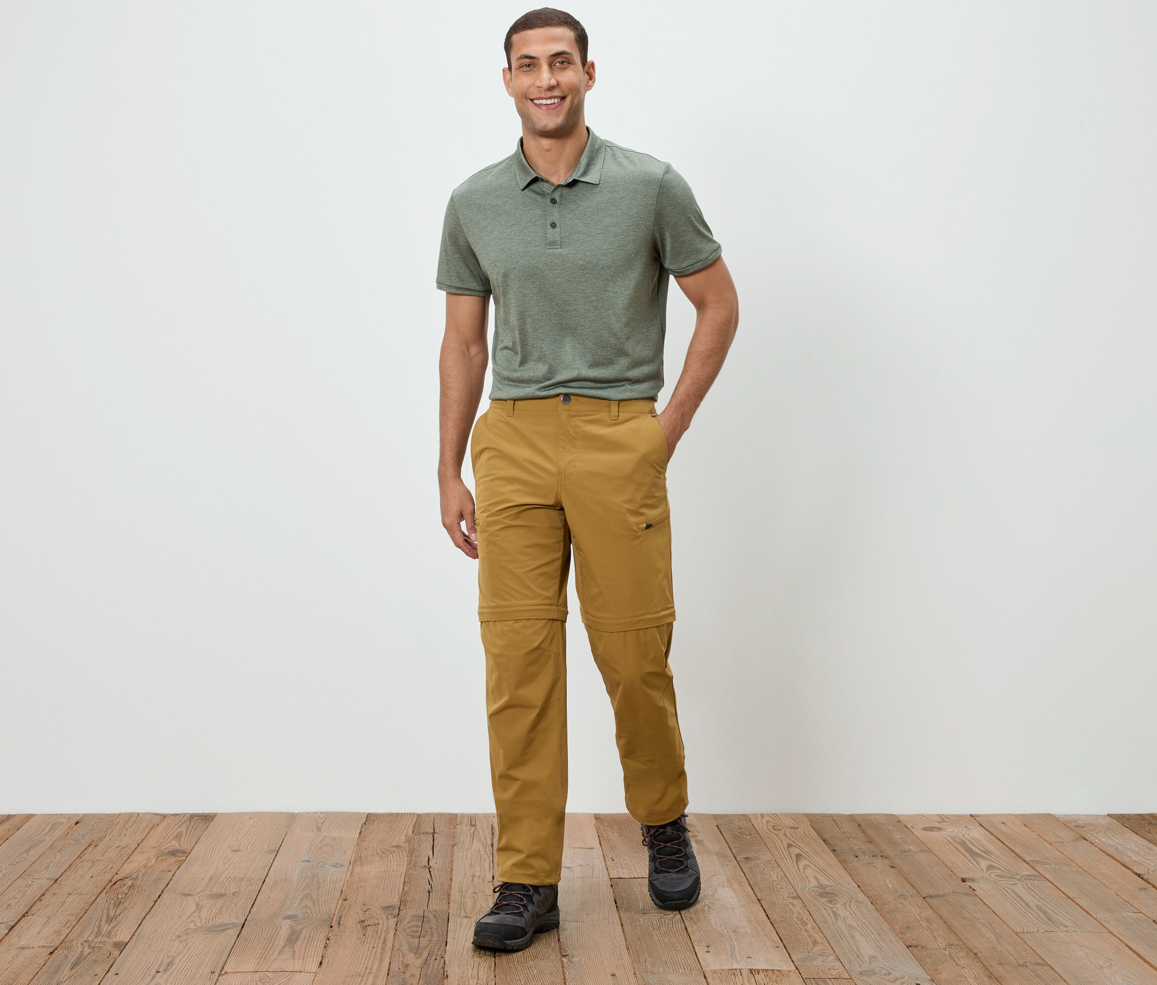 Muž stojí před bílou zdí a má na sobě zelené polo tričko a khaki kalhoty.