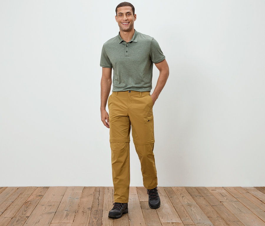 Muž stojí před bílou zdí a má na sobě zelené polo tričko a khaki kalhoty.