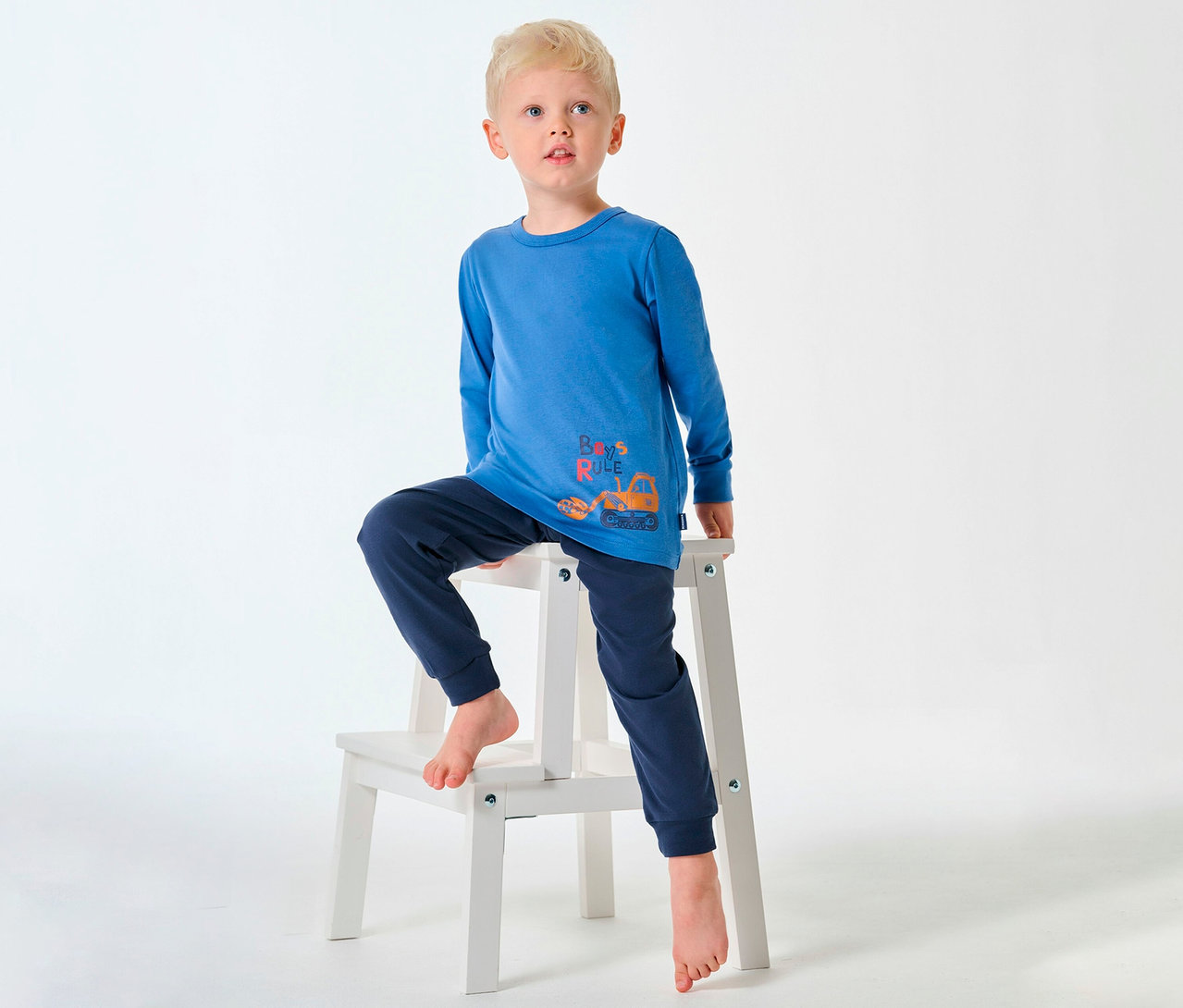 Blonďatý chlapec v dlouhém dětském pyžamu SCHIESSER sedí bosý na stoličce.