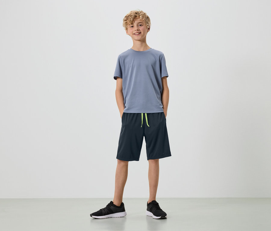 Mladý muž s blond kudrlinami stojí na bílém pozadí v šedém tričku, tmavě modrých šortkách a černých teniskách.