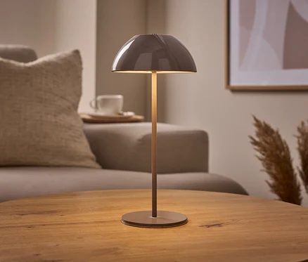Béžová LED dobíjecí stolní lampa s kopulovitým stínítkem stojí na kulatém dřevěném stole. V pozadí je pohovka, šálek a pampová tráva.