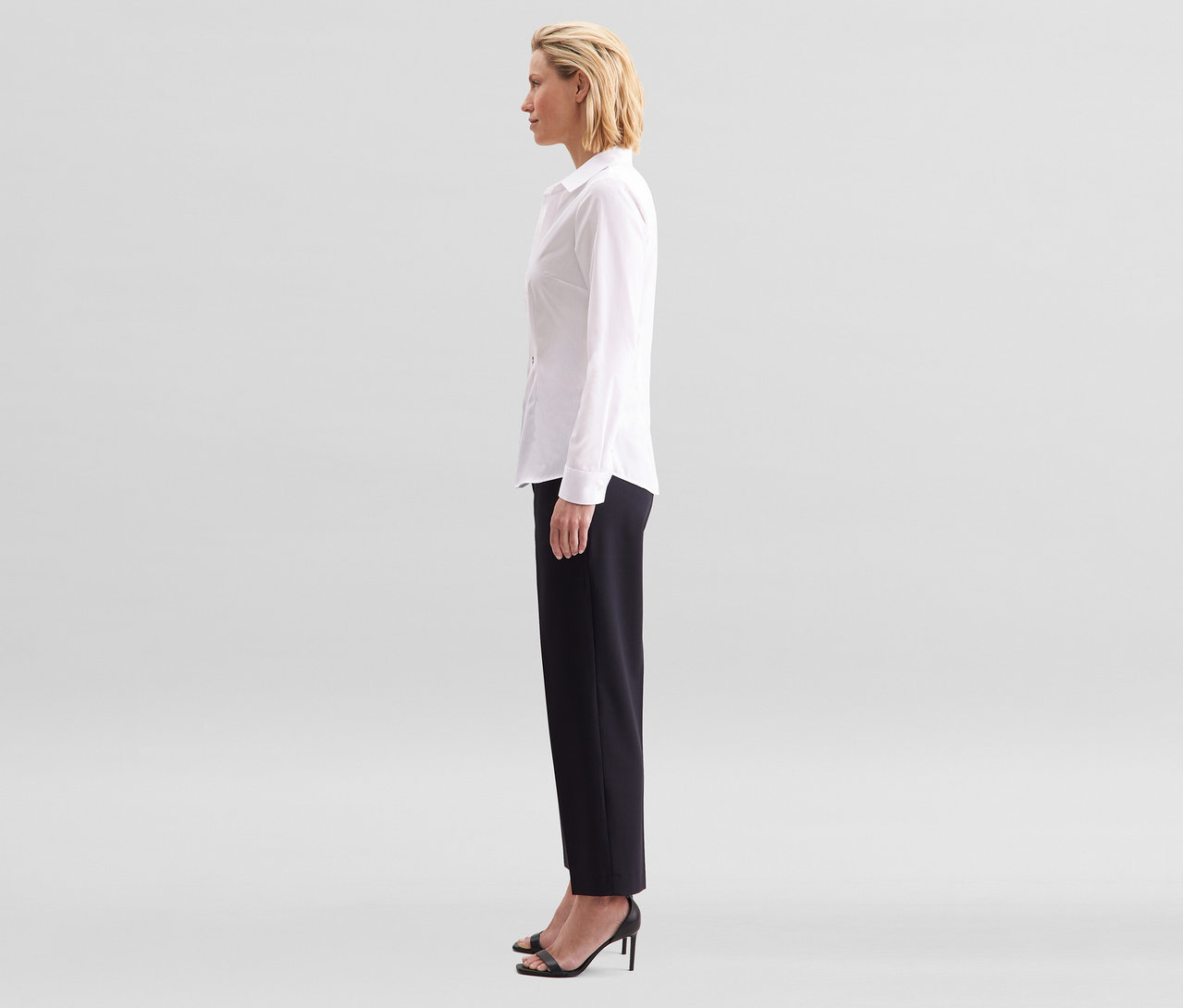 Žena z profilu, oblečená v bílé halence slim fit non iron Seidensticker a černých kalhotách.