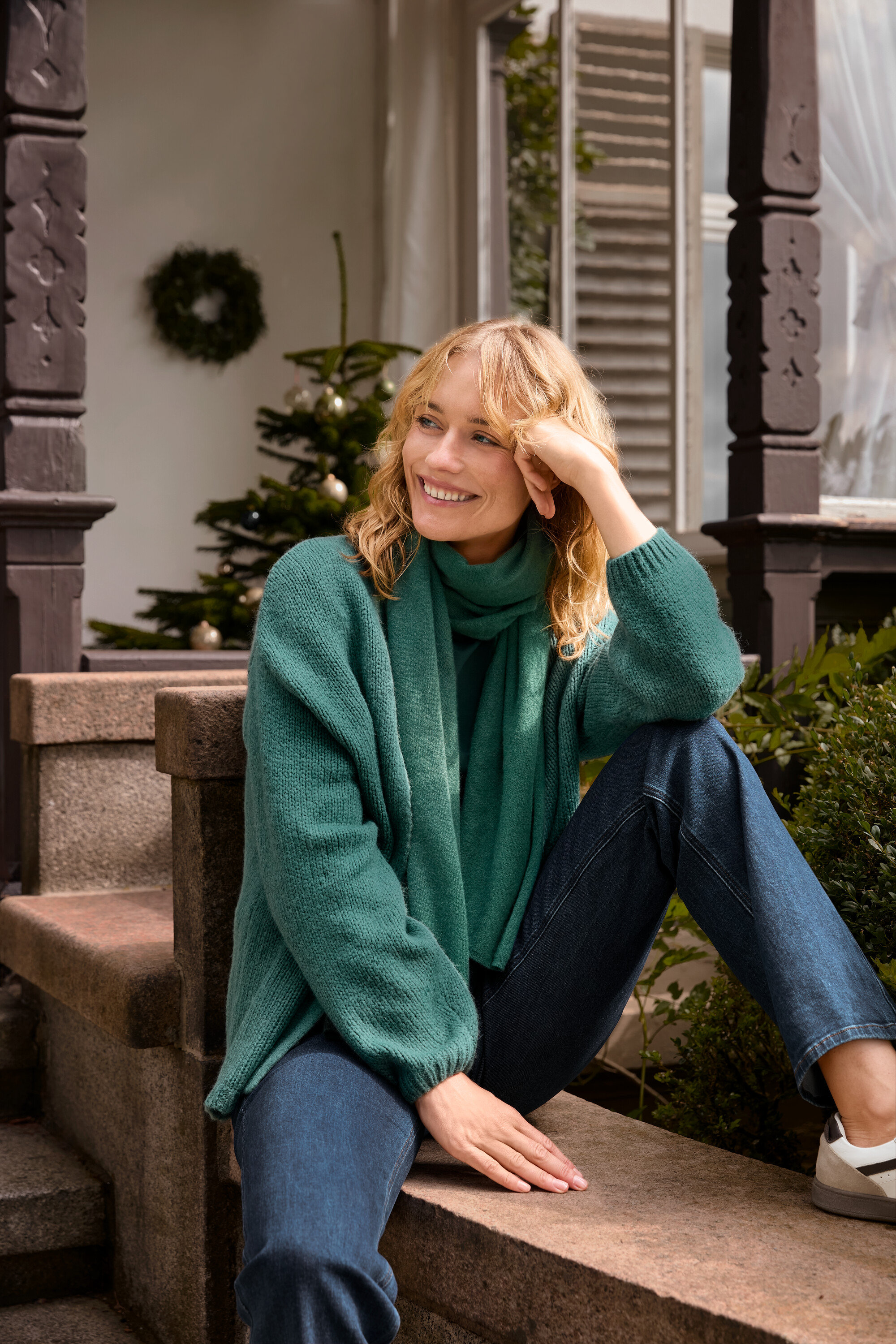 Blonďatá žena sedí na schodech a má na sobě otevřený kardigan z hrubého úpletu, zelenou tkanou šálu a džíny bootcut – Fit »Lou«.