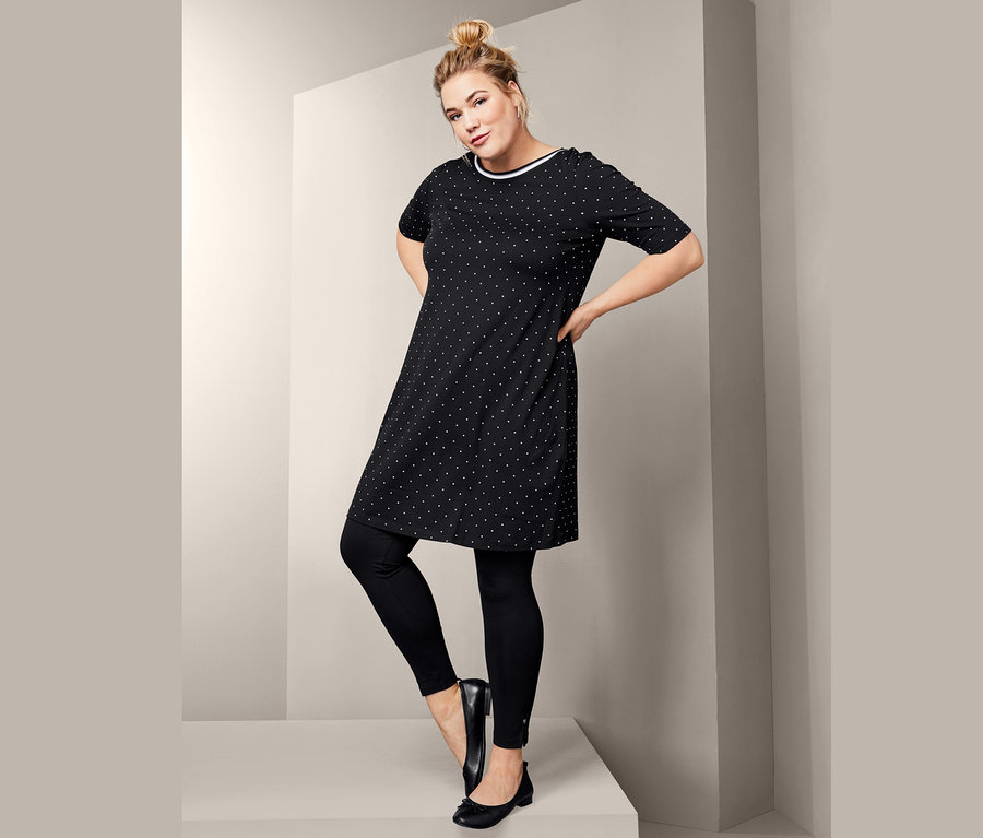 Žena pózuje v černých legínách, kožených balerínách a žerzejových šatech s puntíkovaným vzorem.