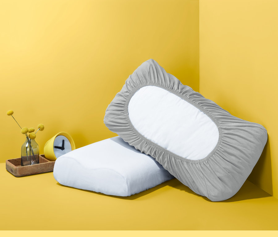 Dva polštáře na žlutém pozadí. Jeden polštář má šedý napínací žerzejový povlak.