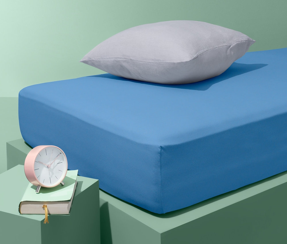 Modré jersey prostěradlo na posteli s šedým polštářem. Na nočním stolku leží růžový budík a kniha.