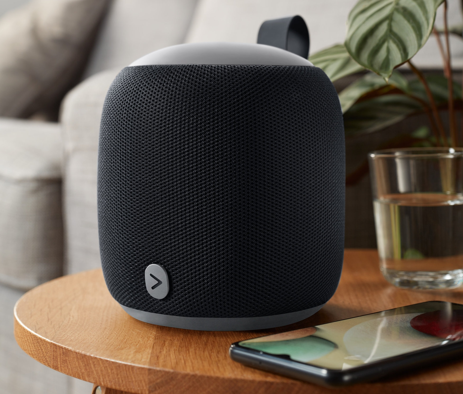 Černý střední Bluetooth® reproduktor Fabric stojí na dřevěném stolku vedle mobilního telefonu.