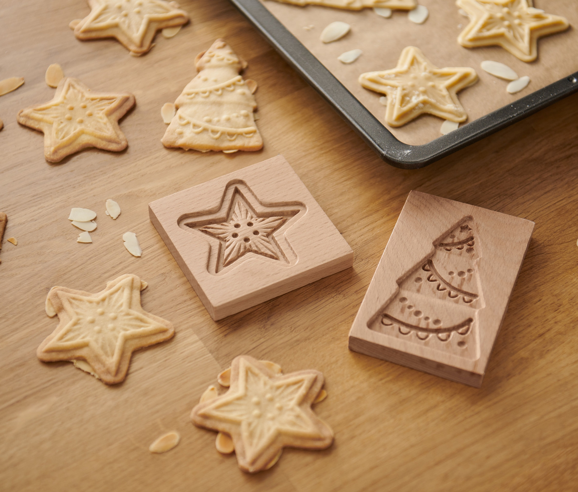 Na dřevěném stole leží formičky na sušenky Spekulatius (2 ks) ve tvaru hvězdy a vánočního stromku spolu s upečenými sušenkami a mandlovými lupínky.
