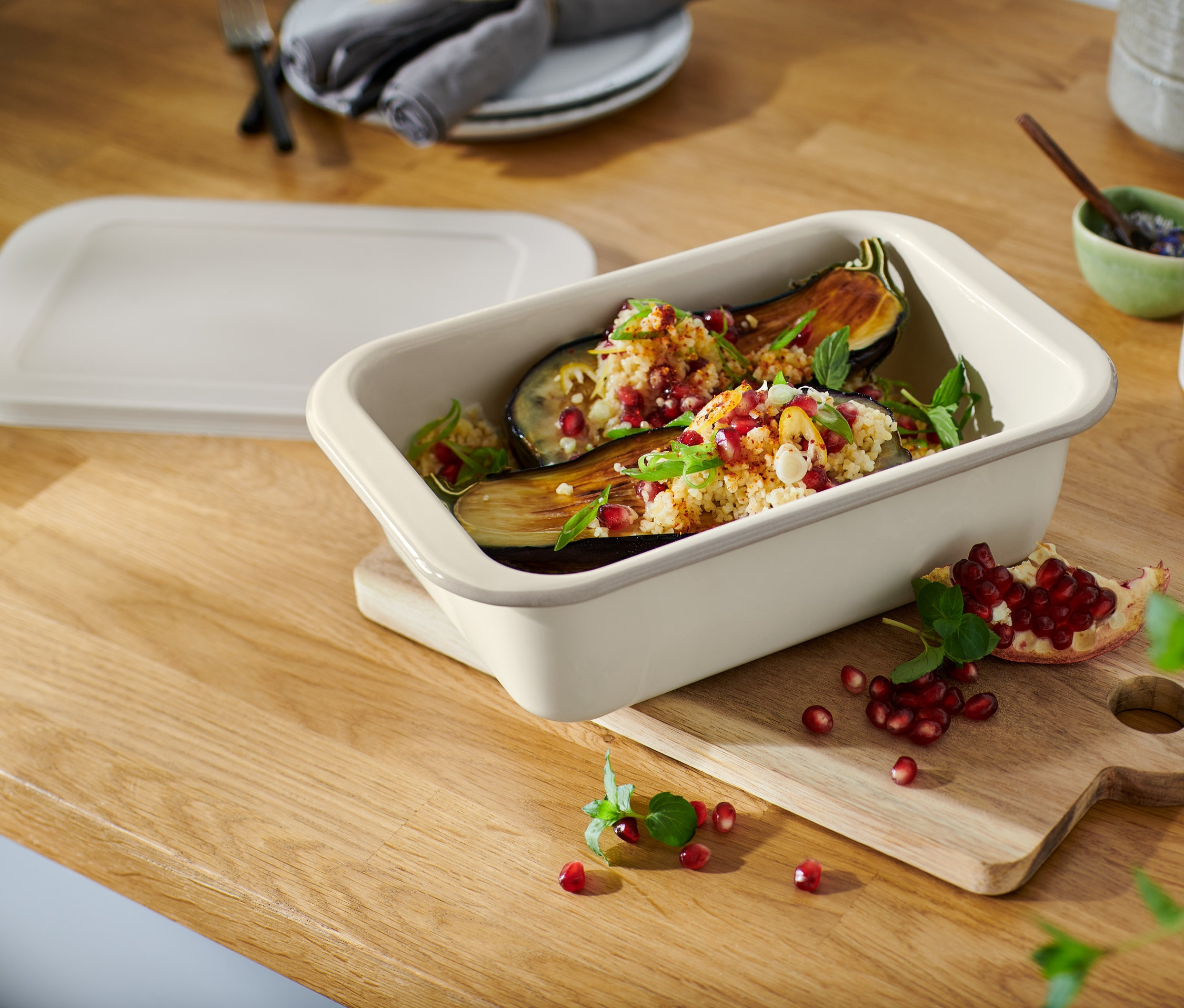 Pečené lilky v malé smaltované zapékací formě s víkem, posypané granátovým jablkem, na dřevěném prkénku.