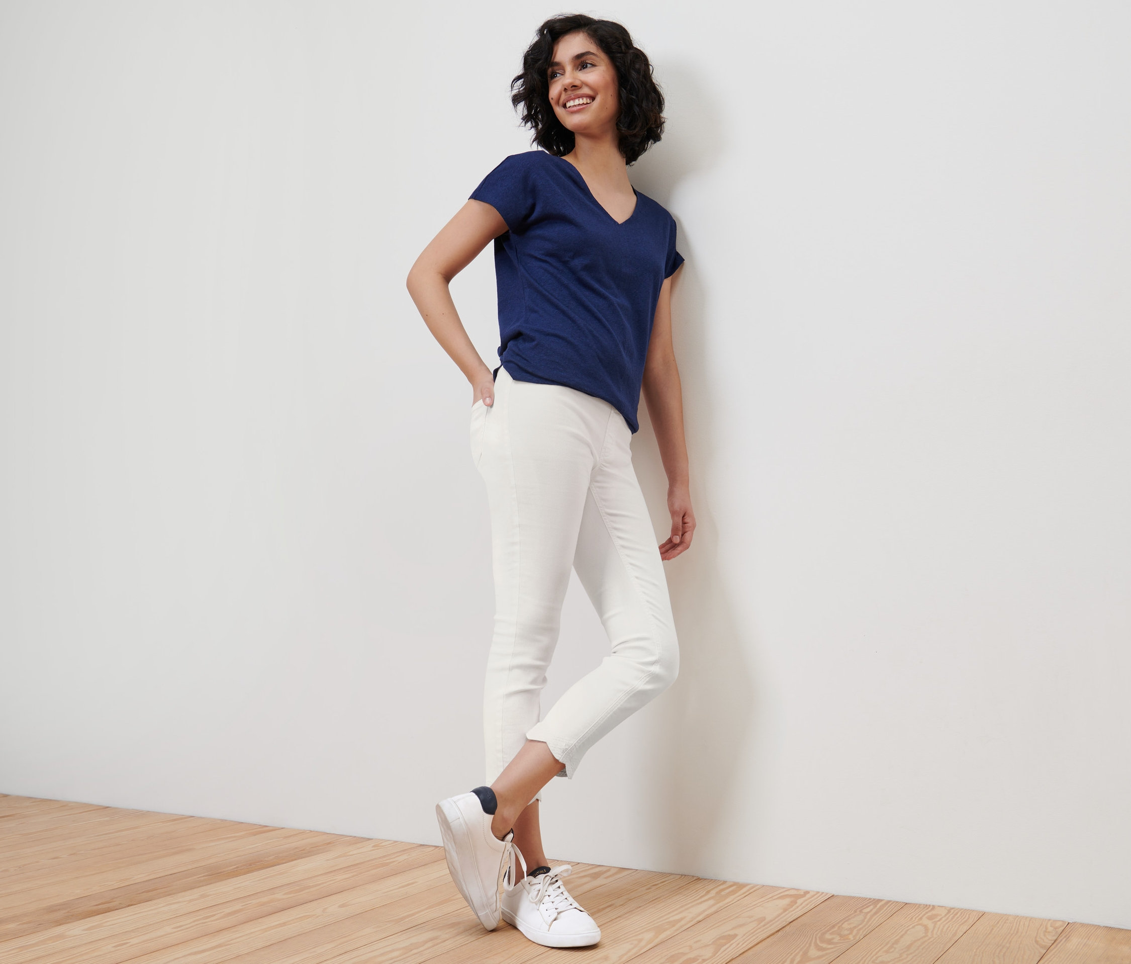 Usmívající se žena opřená o zeď má na sobě modré tričko a bílé 7/8 tregíny s ažurovou výšivkou.