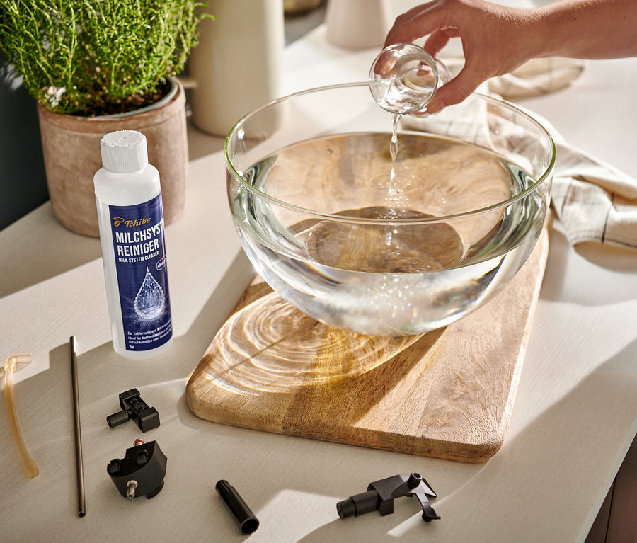 Ruka nalévá čirou tekutinu z odměrky do skleněné mísy s vodou. Vedle mísy stojí čistič mléčného systému durgol® a součástky kávovaru.