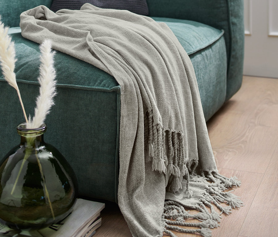 Šedá žinylková deka s třásněmi přehozená přes zelenou pohovku.