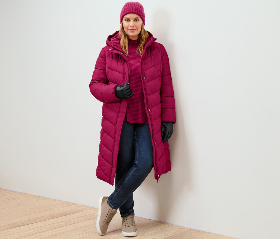Žena má na sobě prošívaný kabát s kapucí malinové barvy, džíny slim – Fit »Emma« tmavě modré a halenkové triko se stojáčkem a potiskem.