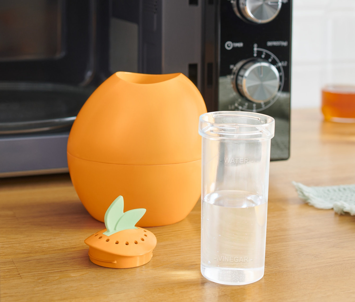 Oranžový čistič do mikrovlnné trouby vedle sklenice s vodou a mikrovlnné trouby.