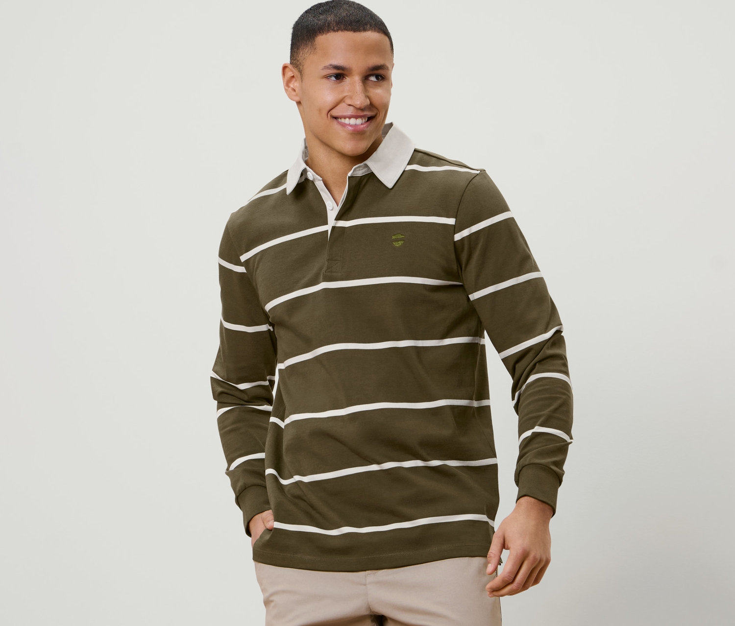 Muž s krátkými vlasy usmívá se a pózuje v olivově zelené polokošili s dlouhým rukávem a bílými pruhy.