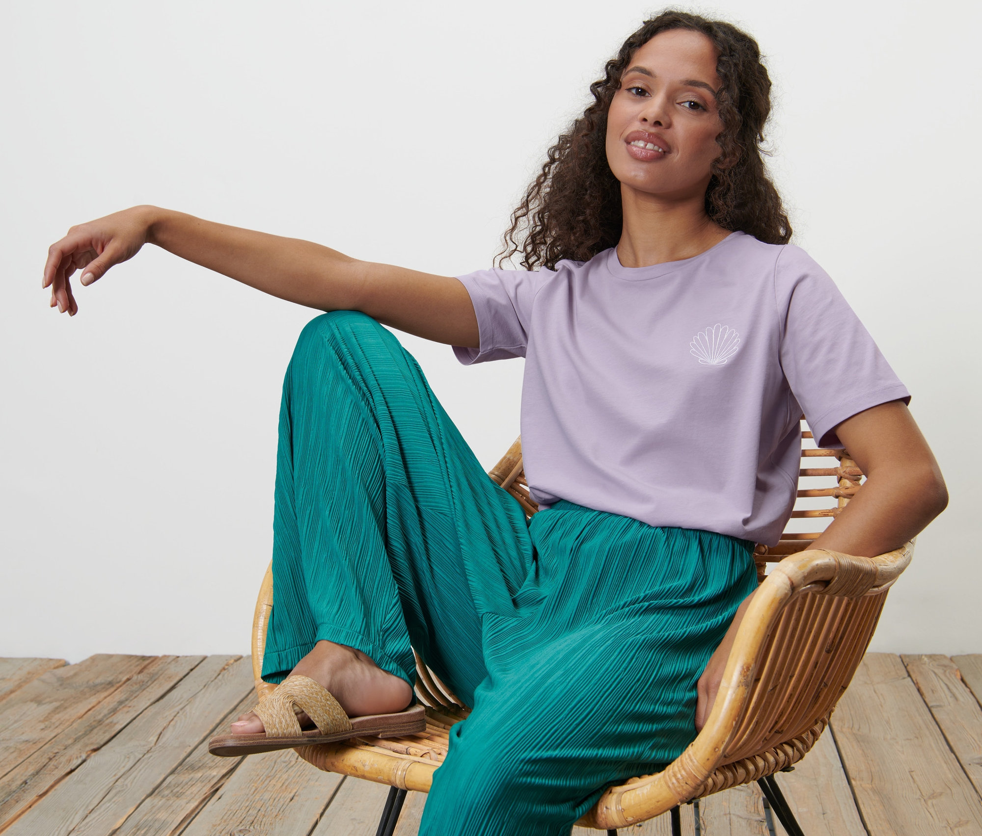 Žena sedí v proutěném křesle a má na sobě fialové tričko s výšivkou a zelené kalhoty.