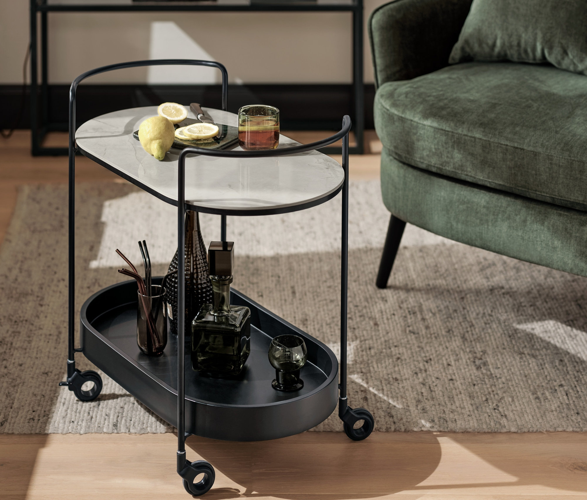 Černý servírovací vozík s mramorovou deskou, nápoji a sklenicemi. V pozadí je konzole v mramorovém vzhledu a stolní lampa se stínítkem z kouřového skla.