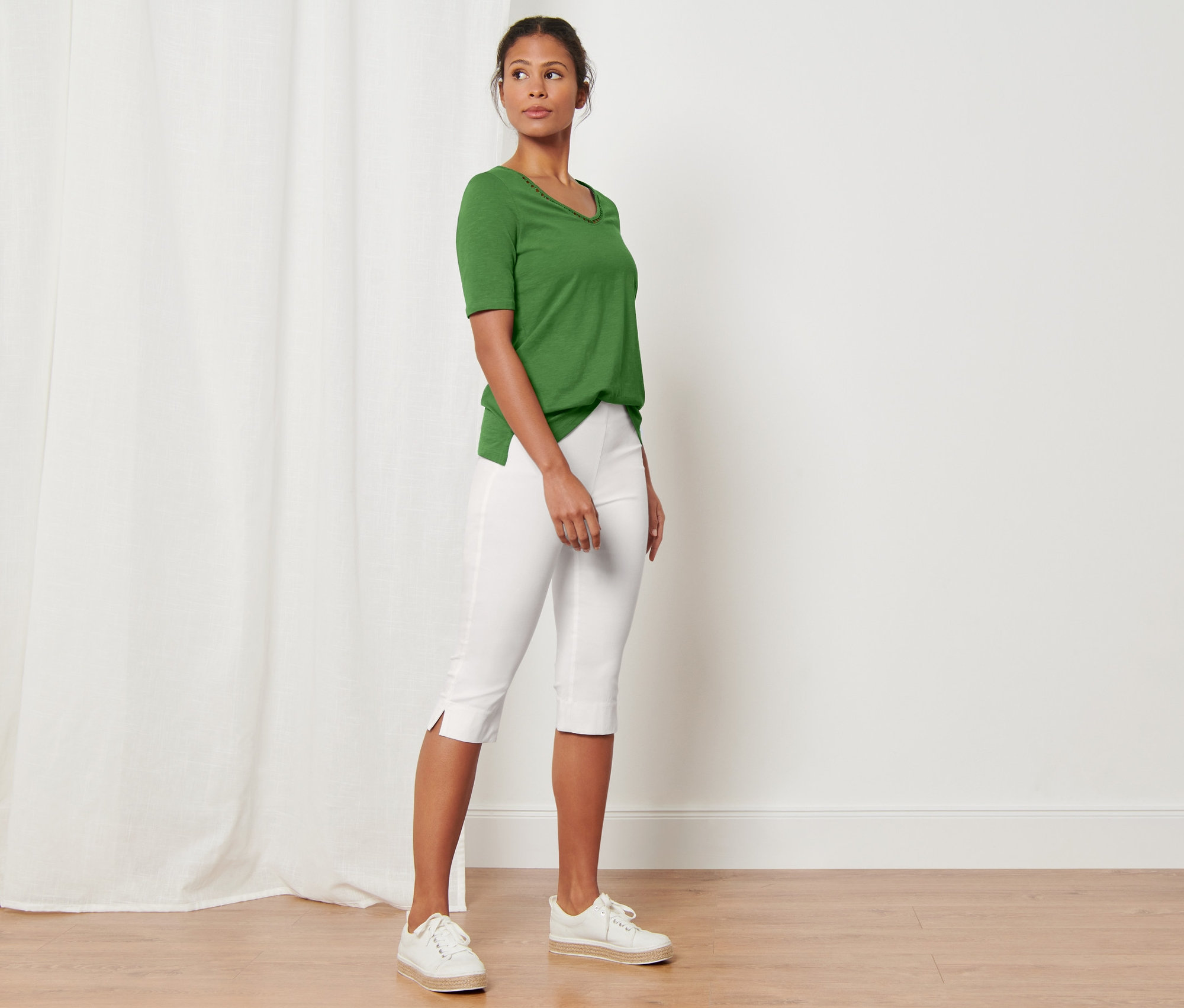 Žena stojící v místnosti má na sobě zelené tričko a bílé 3/4 strečové kalhoty.