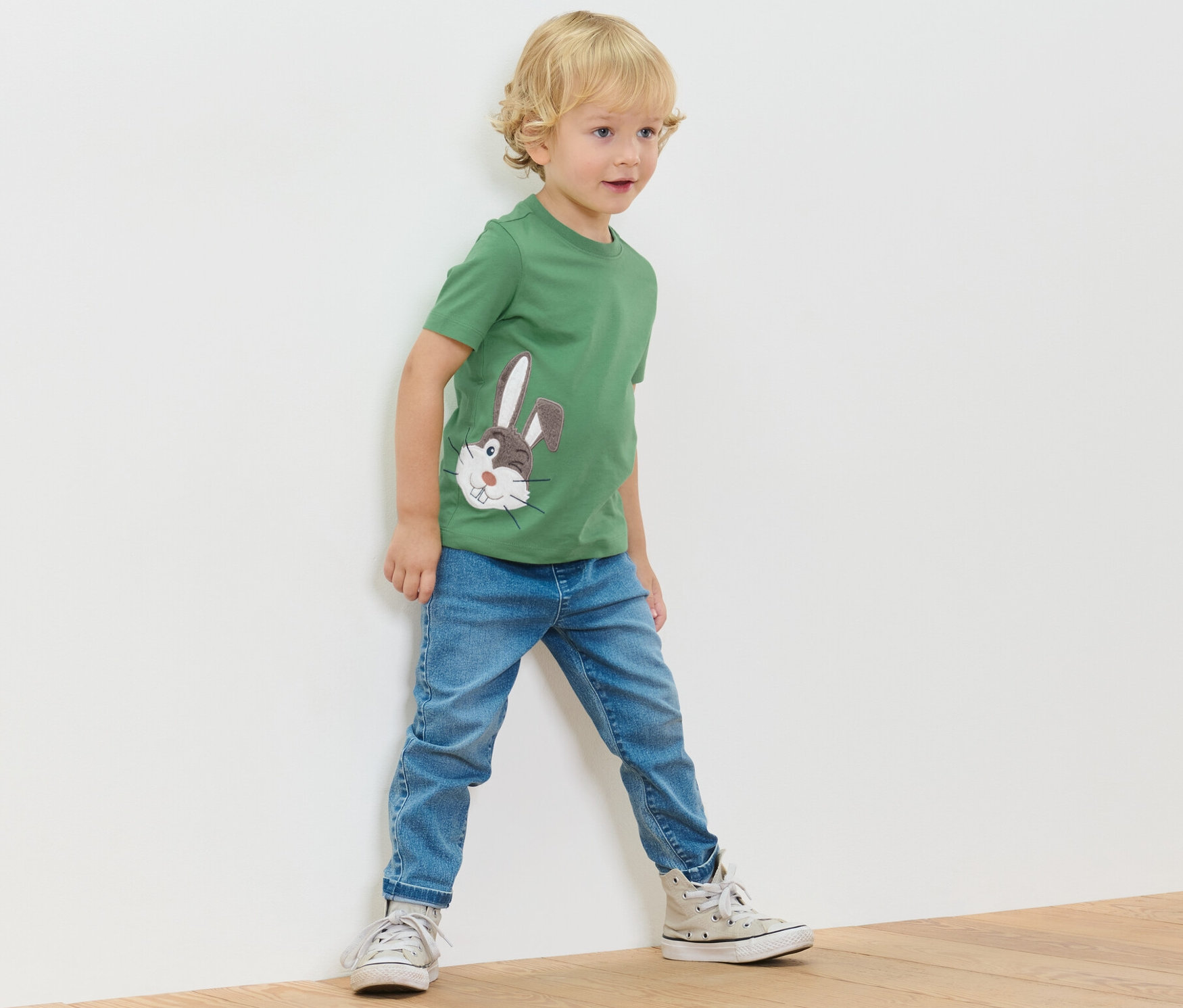 Chlapec stojí u stěny a má na sobě zelené tričko s aplikací a džíny.