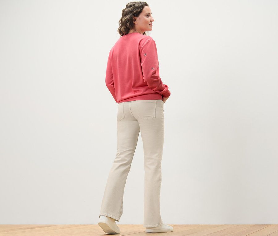 Žena stojí zády k fotoaparátu a má na sobě krémově bílé džíny bootcut střihu »Lou« a růžovou mikinu.