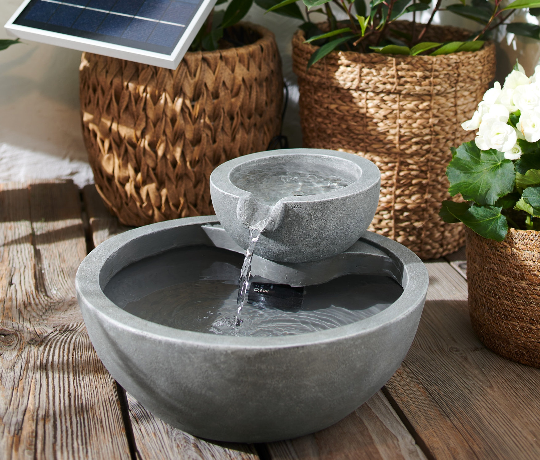 Tekoucí solární zahradní fontána na dřevěné podlaze, v pozadí rostliny v proutěných koších.