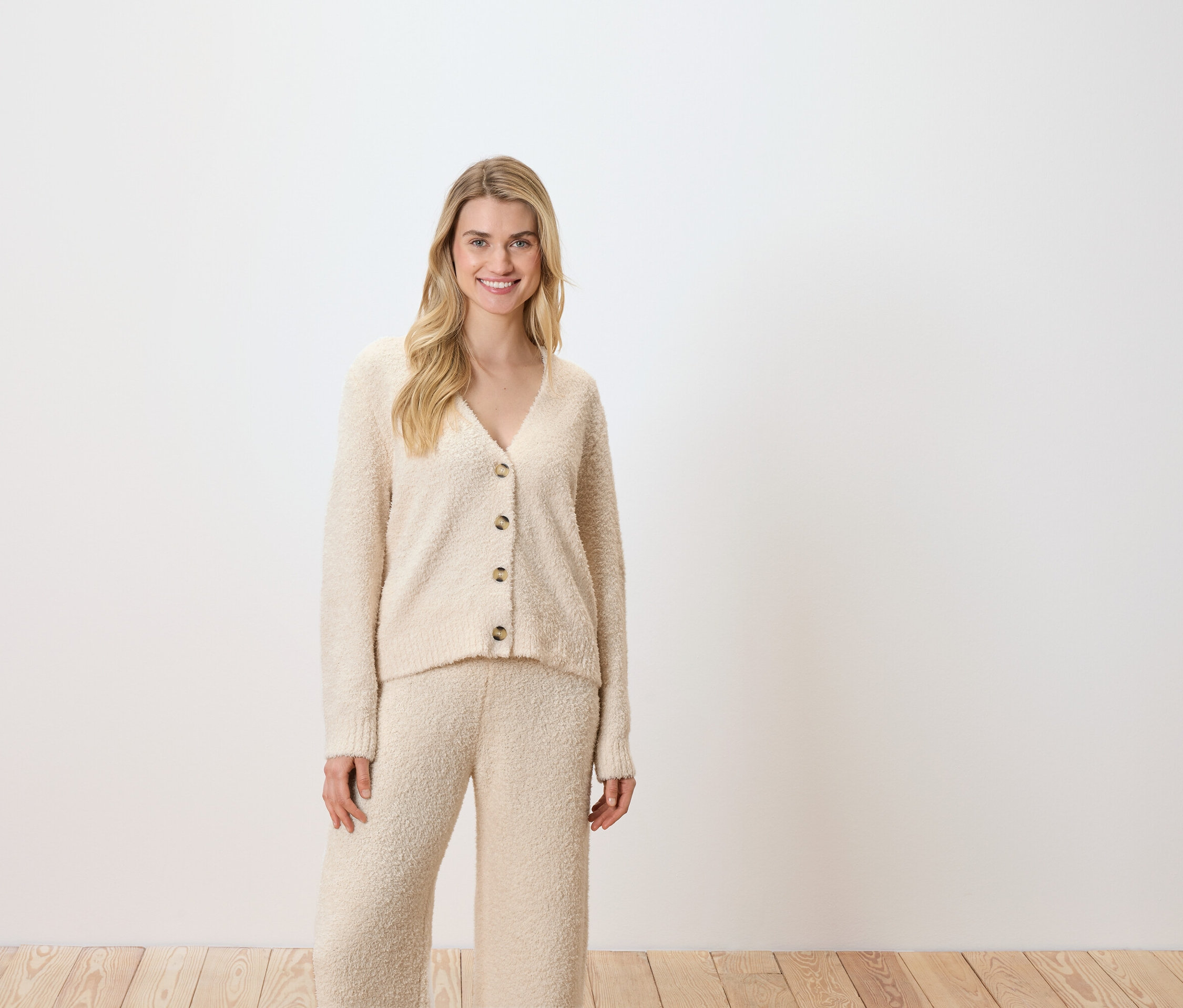 Žena pózuje na bílém pozadí a má na sobě krémový kardigan a kalhoty.