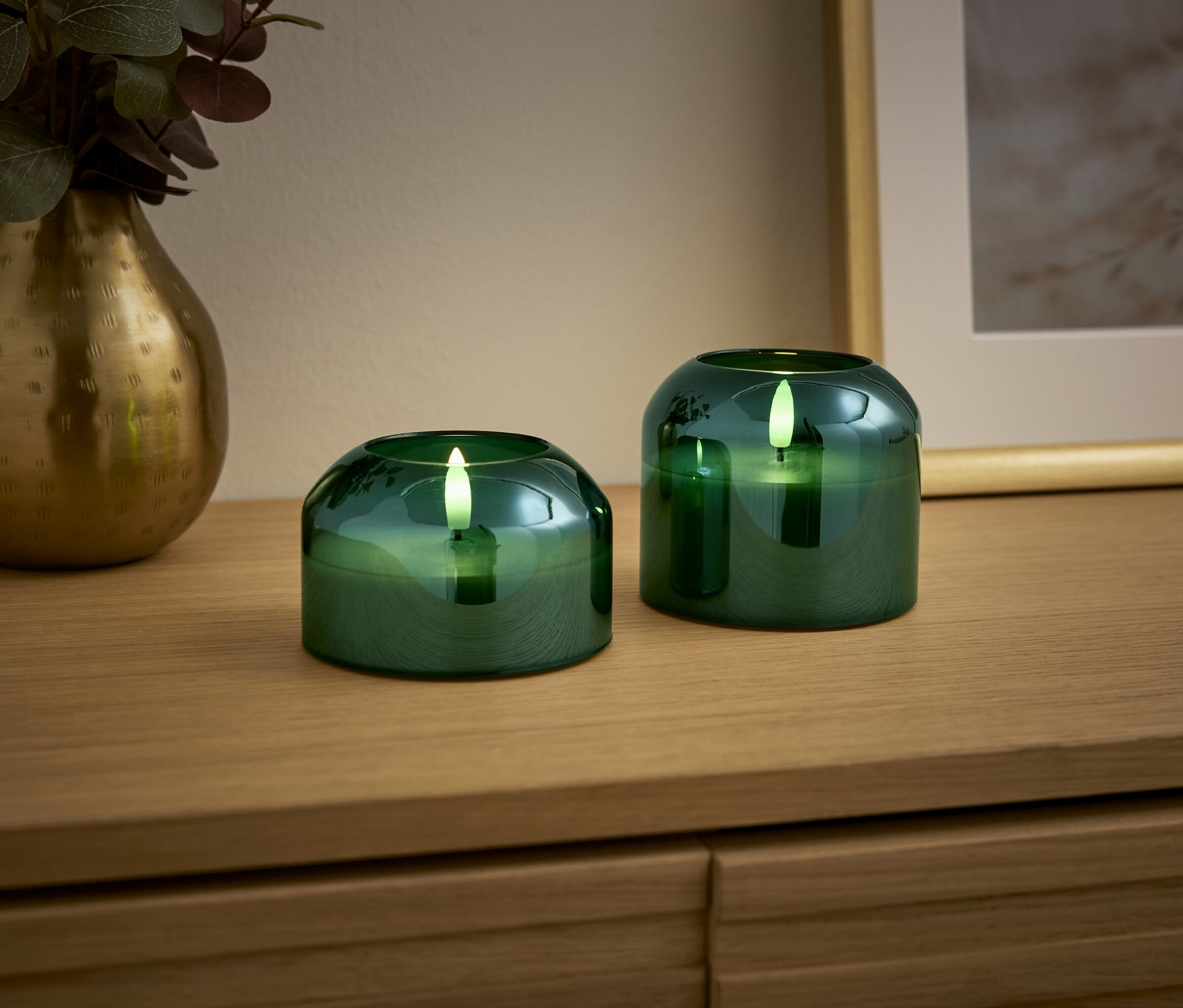 Dvě zelené svíčky a zlatá váza stojí na dřevěném stole.
