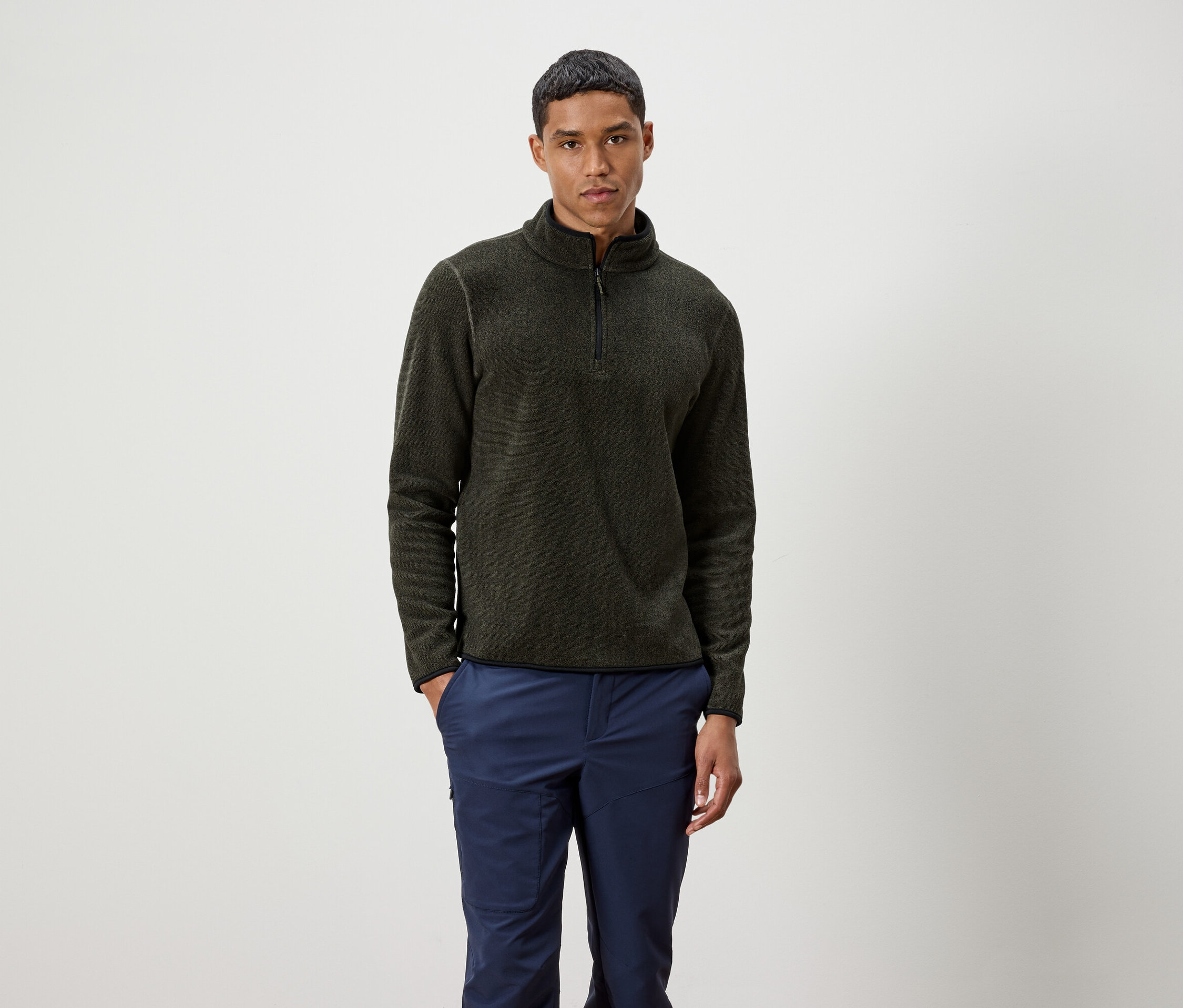 Muž stojí a má na sobě olivový svetr na zip a modré kalhoty.