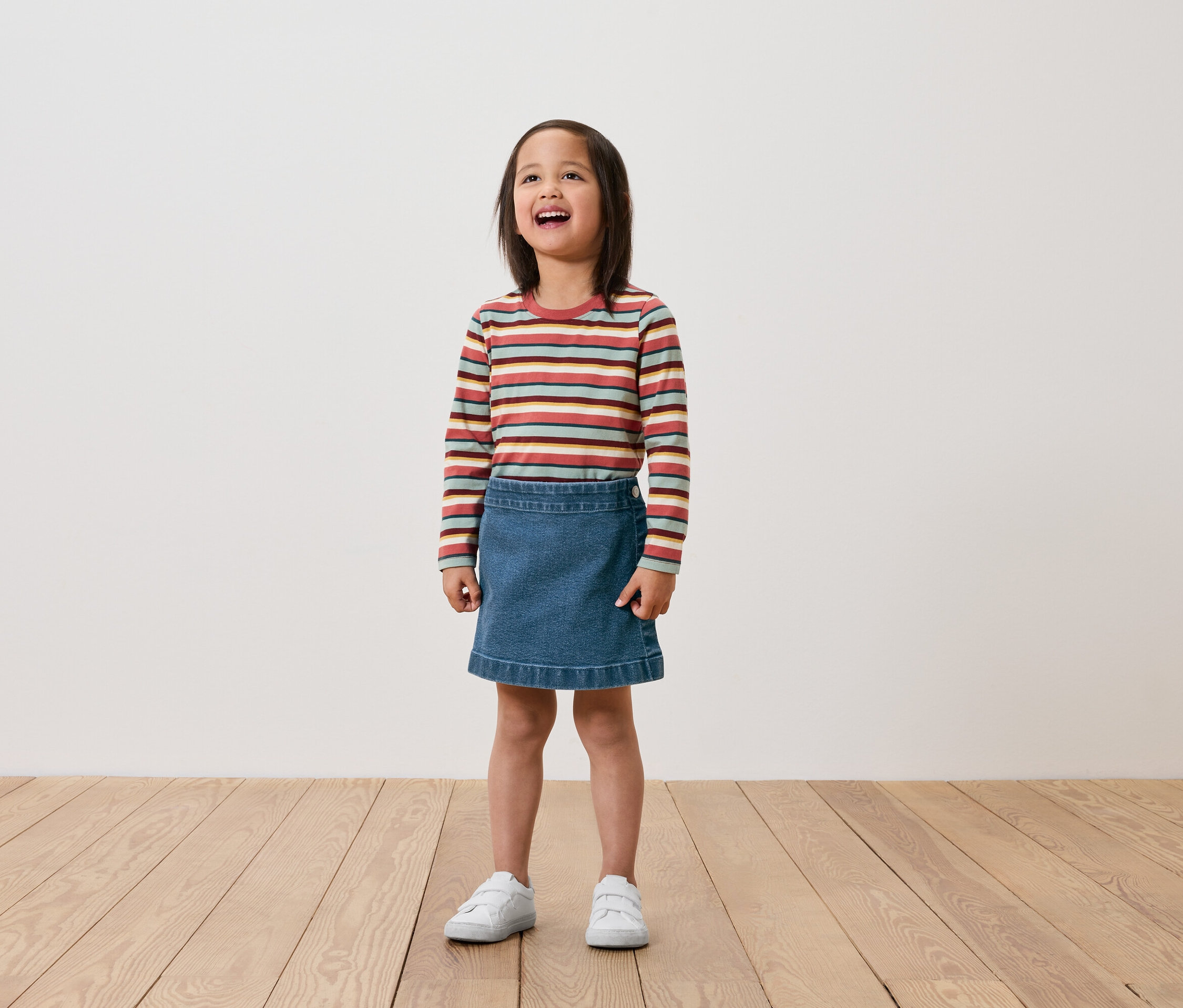 Dívka stojí na dřevěné podlaze a má na sobě pruhované tričko s dlouhým rukávem, džínovou sukni a bílé tenisky.