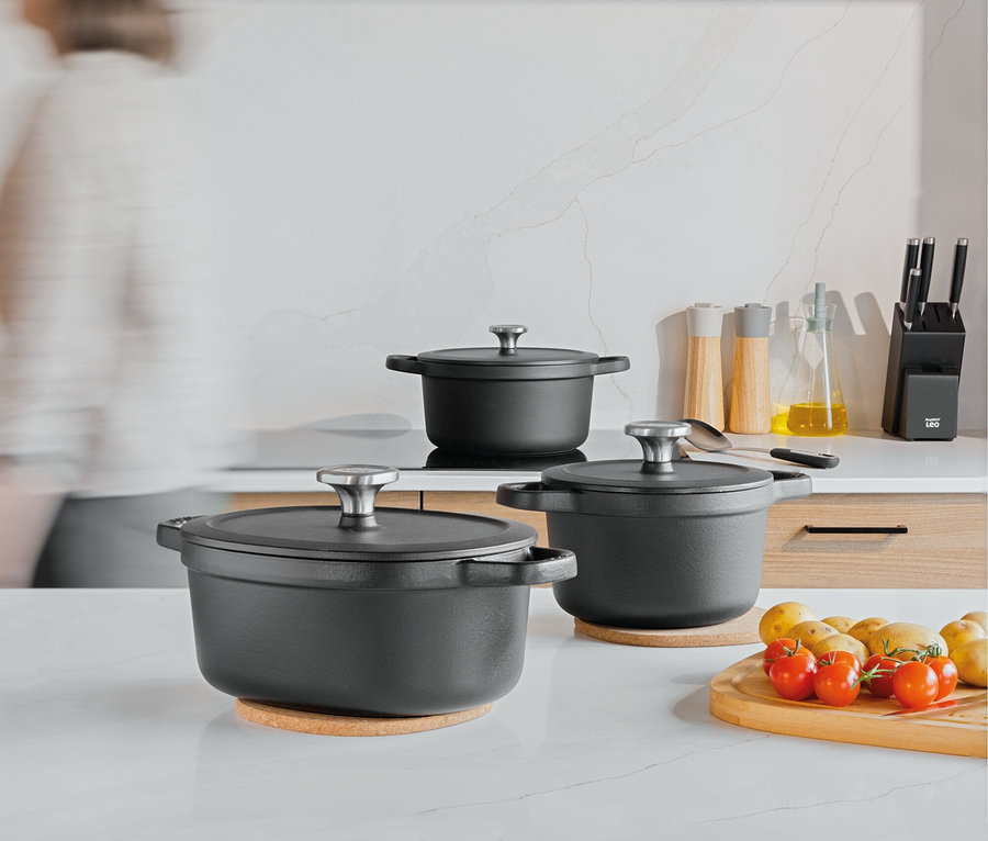 Tři černé hrnce s poklicemi na kuchyňské lince, vedle nich prkénko s rajčaty a bramborami.