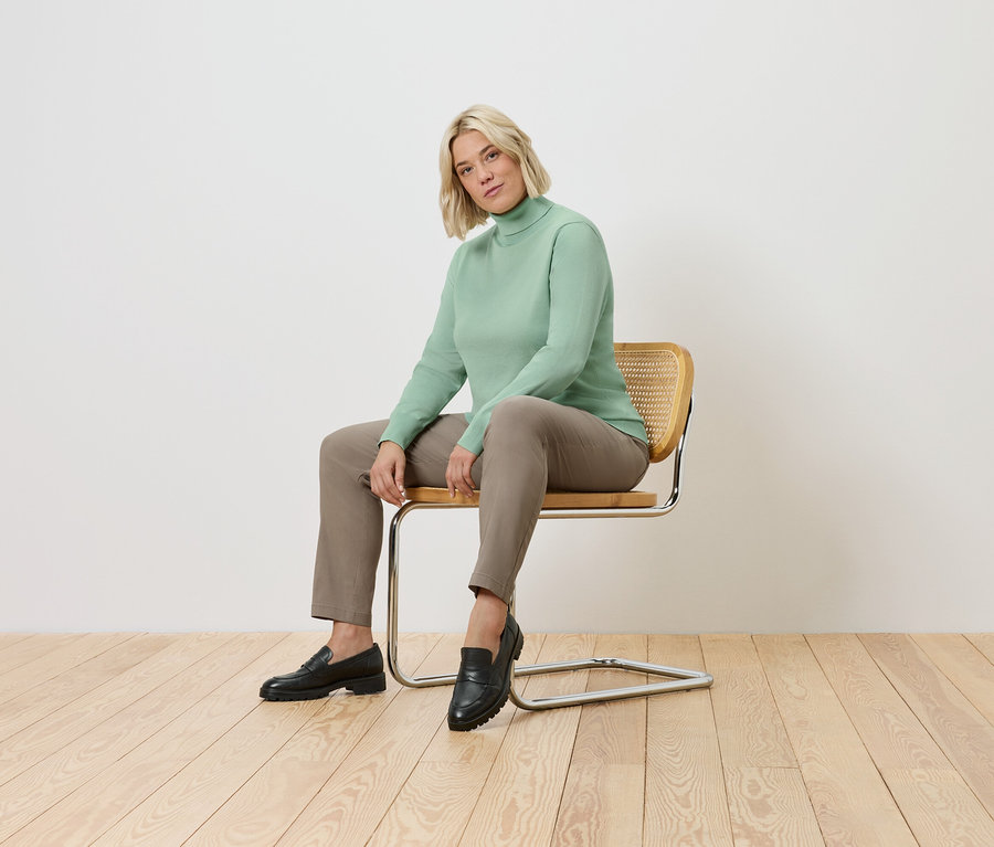 Žena sedí na židli. Má na sobě zelený rolák, hnědé kalhoty a černé boty.