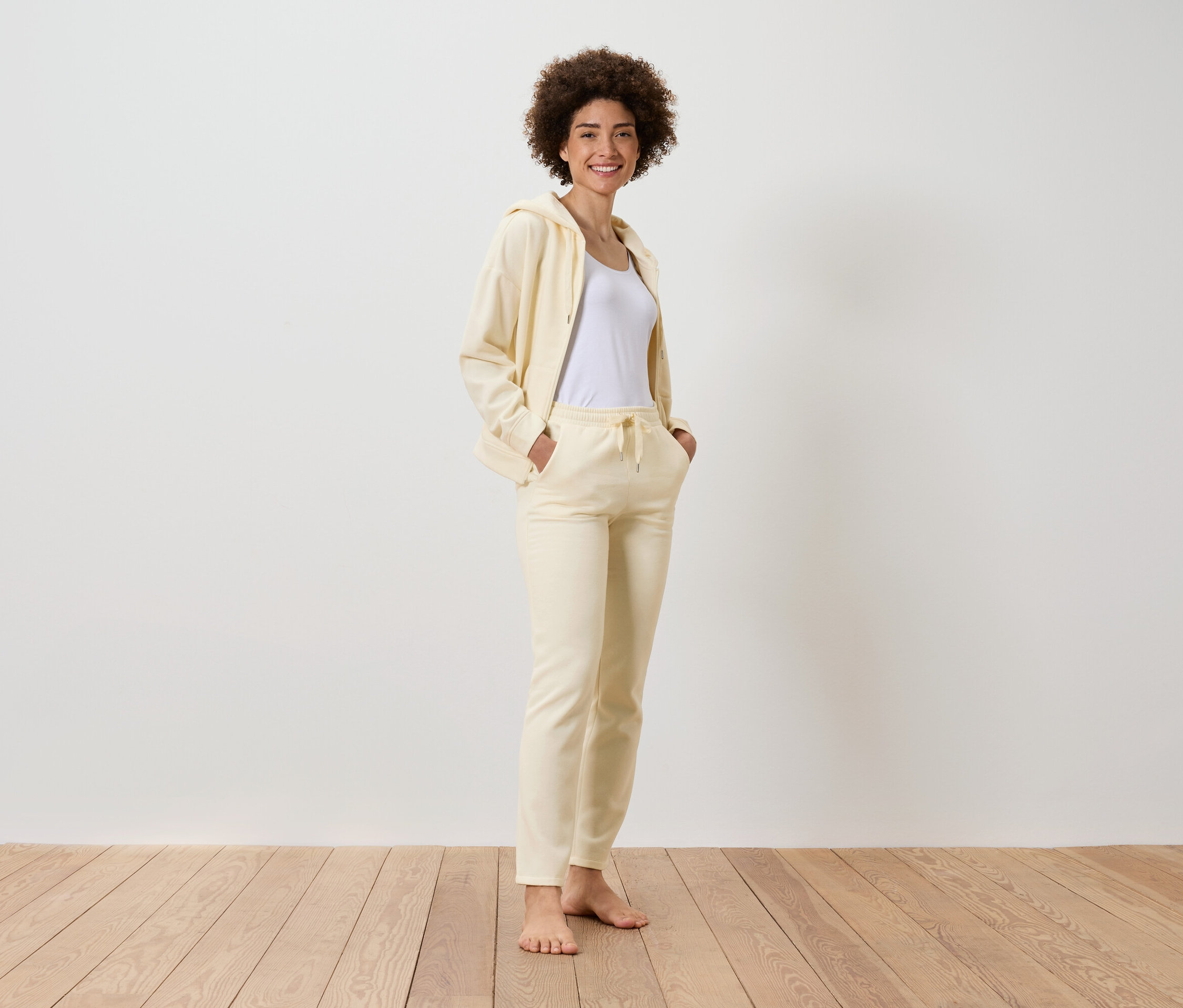 Usmívající se žena s afro účesem stojí a má na sobě žlutou teplákovou soupravu s bílým tričkem.