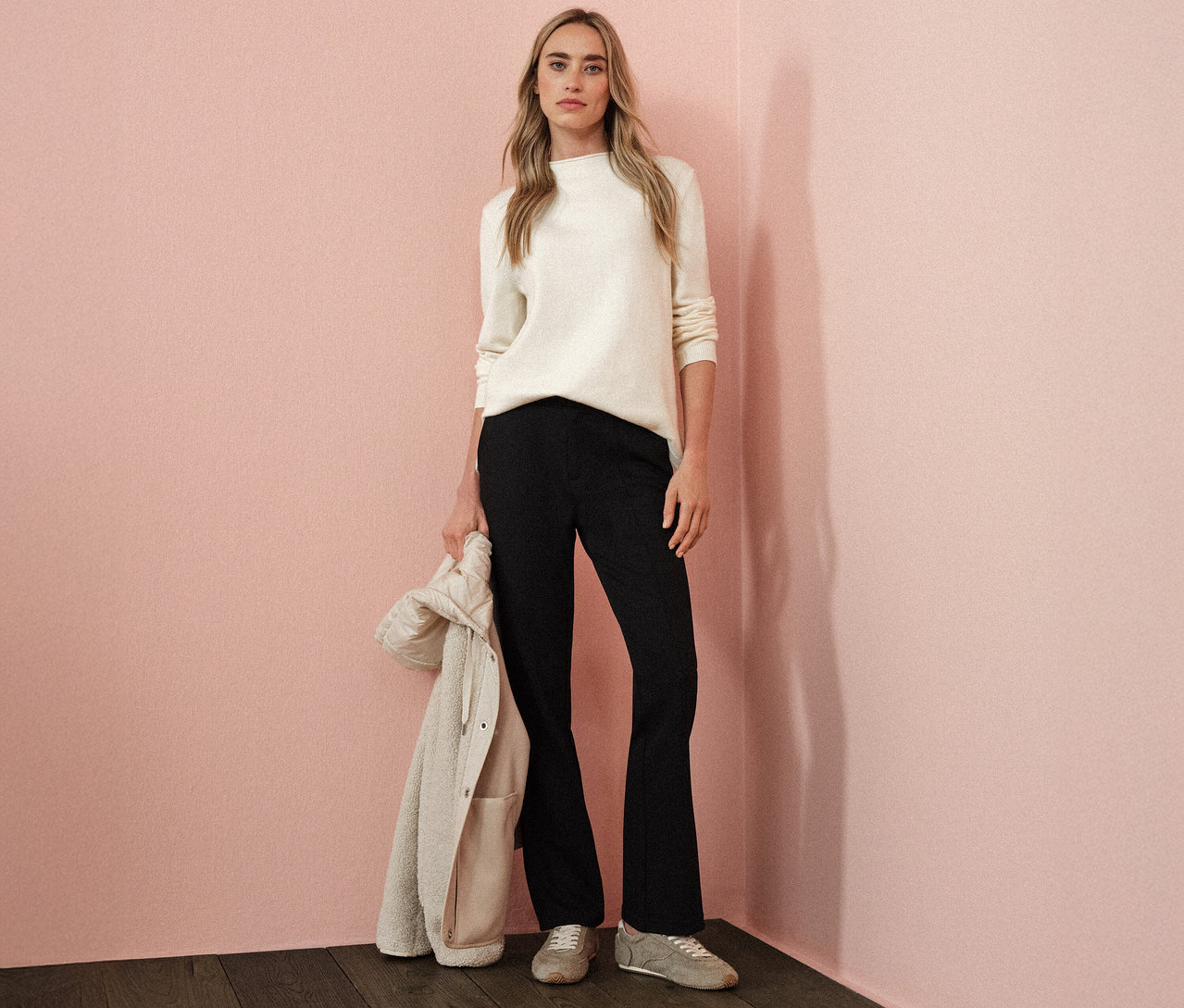 Žena má na sobě béžový pletený svetr s rolovaným lemem Street One, černé kalhoty a drží bundu.