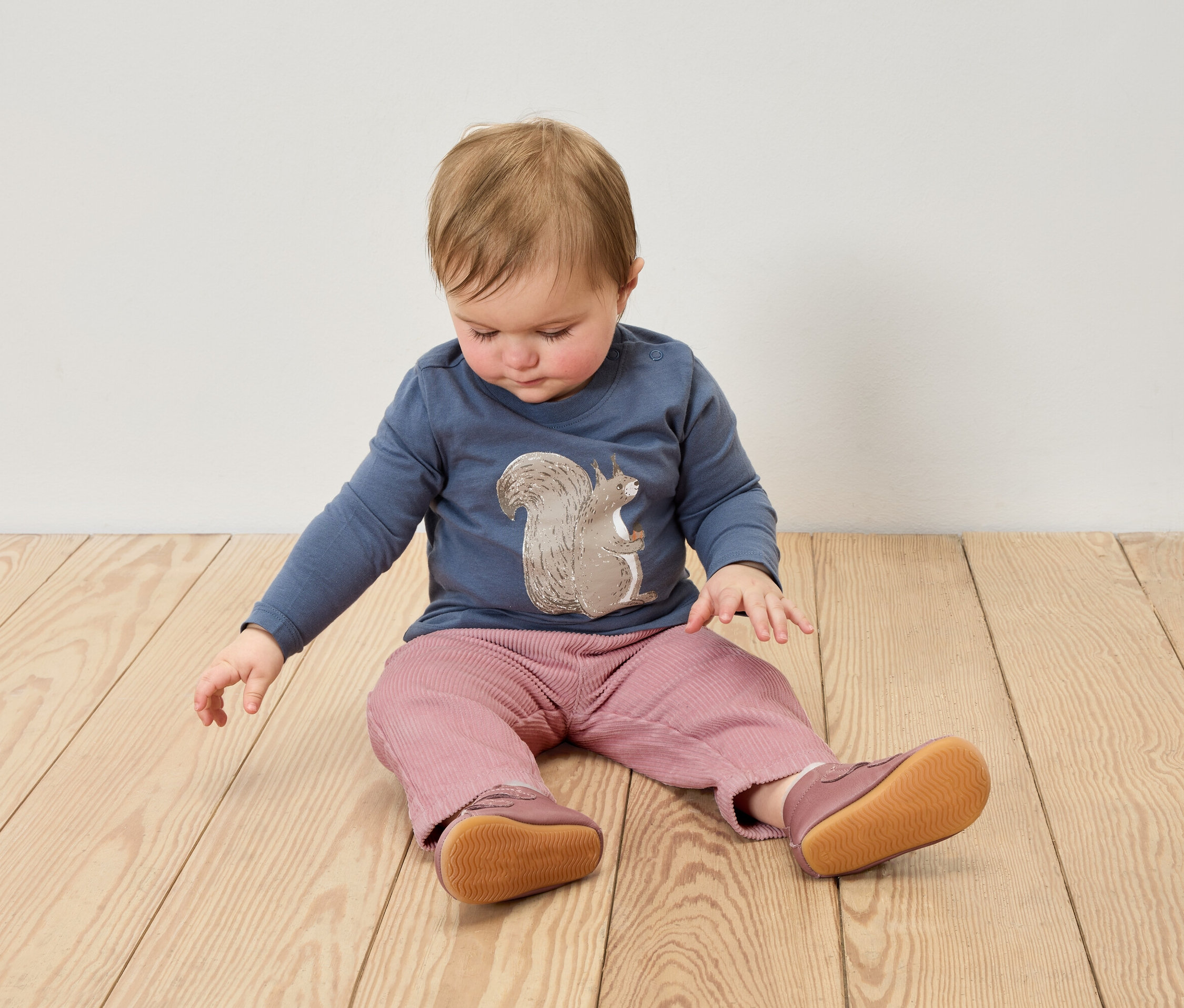 Batole sedí na dřevěné podlaze, má na sobě modré tričko s potiskem veverky, růžové kalhoty a kožené capáčky.