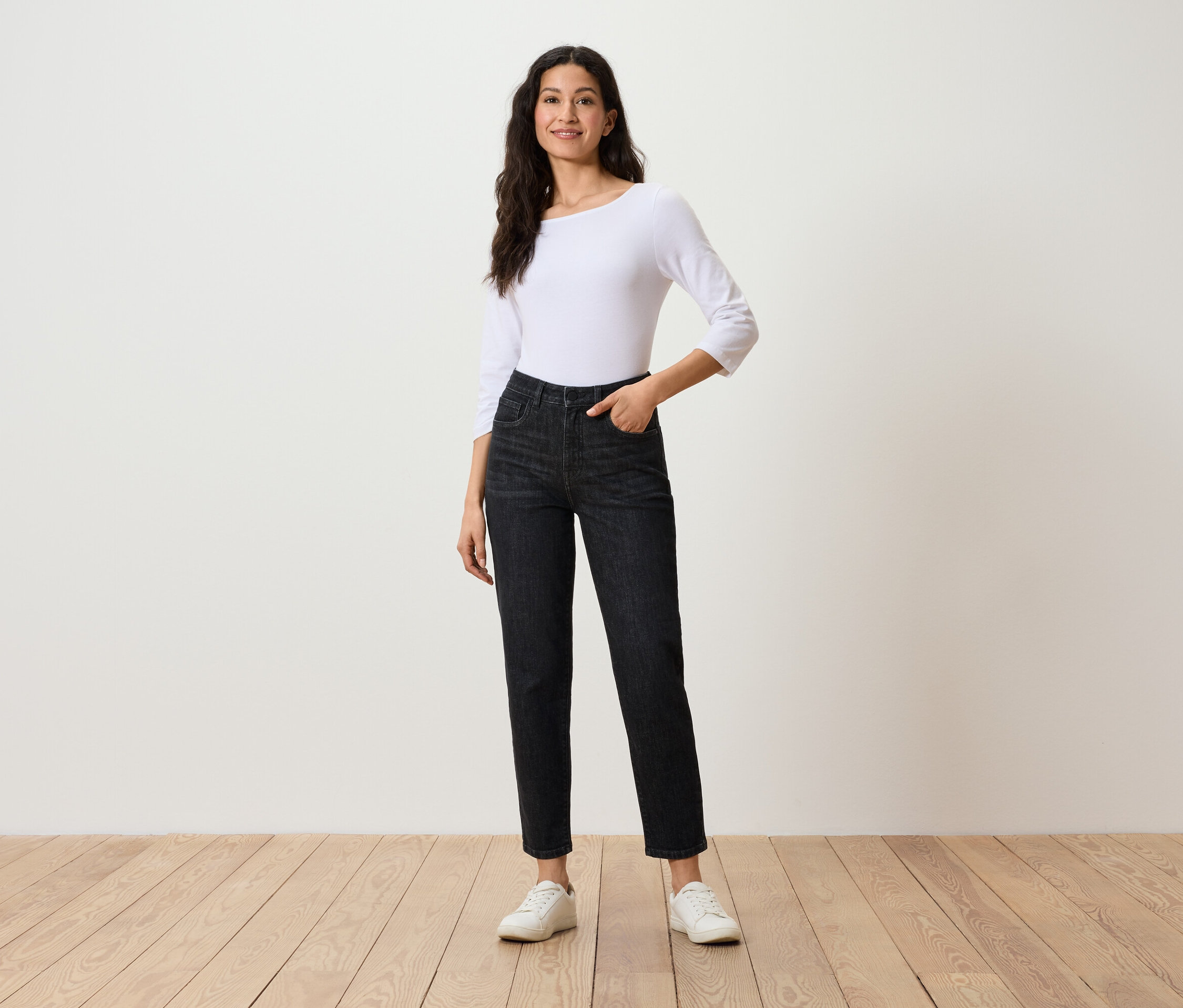 Žena pózuje v černých džínách, bílém topu s tříčtvrtečními rukávy a bílých teniskách.