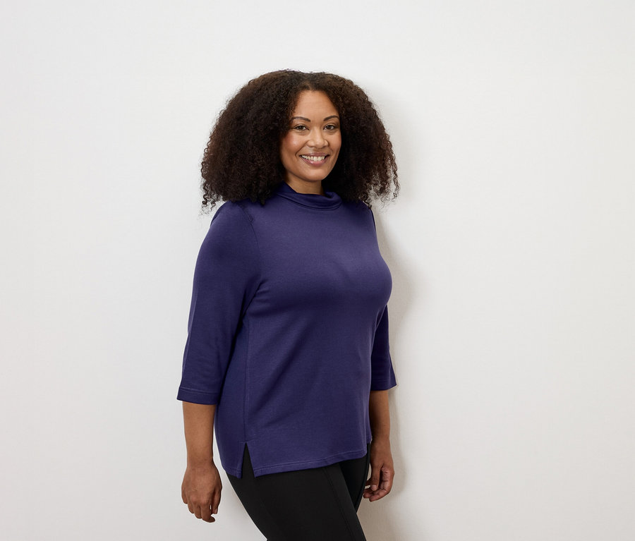 Žena má na sobě lilkový svetr se stojáčkem a pevné legíny s lemovkou.