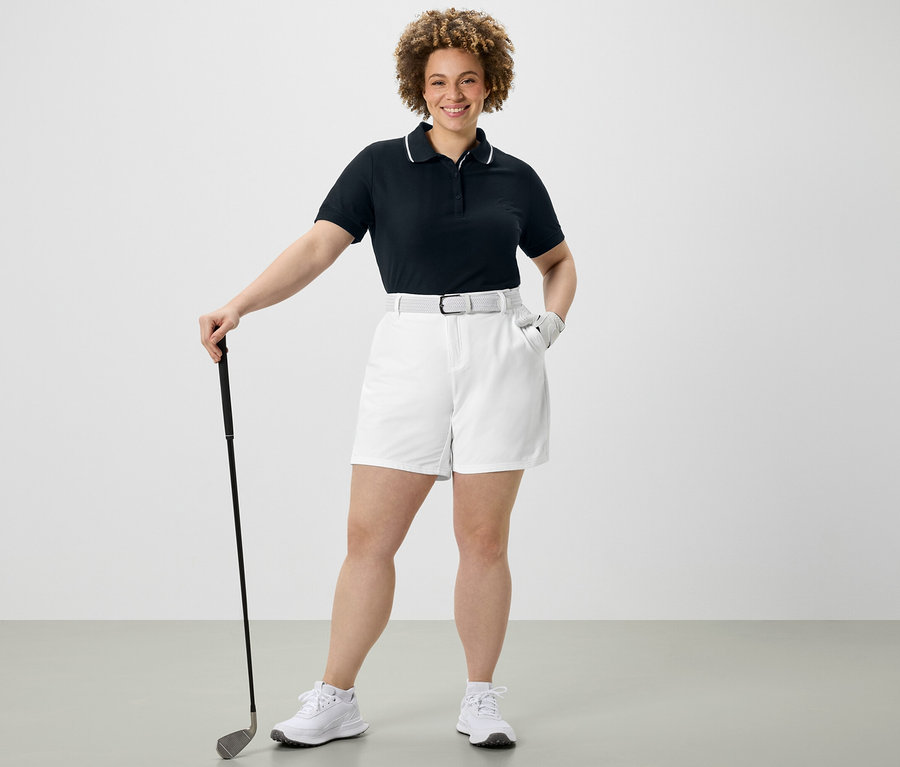 Žena drží golfovou hůl, má na sobě tmavě modré funkční polo triko a bílé softshellové funkční šortky.