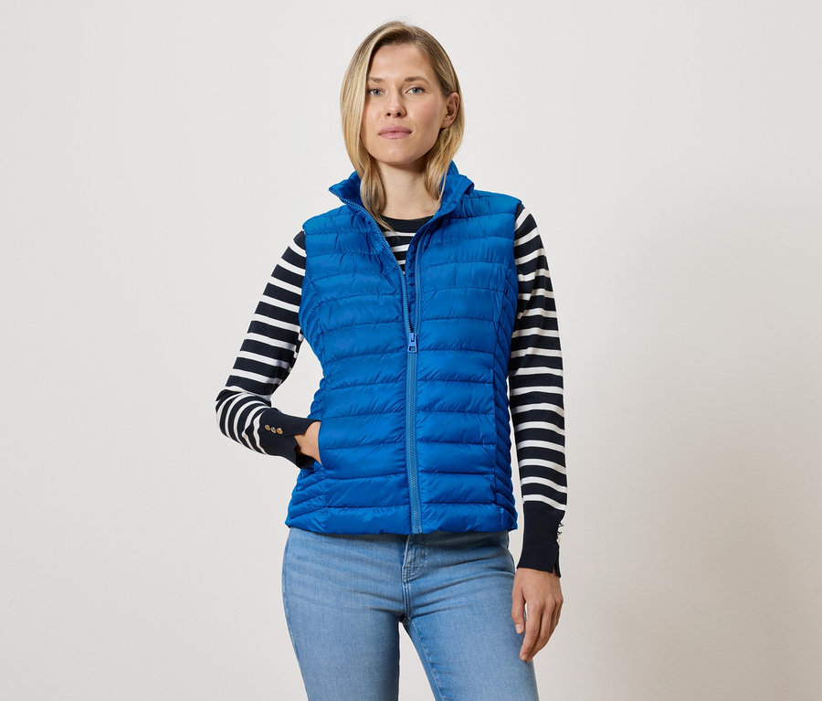 Žena v modré prošívané vestě se zipem, svetru s černými a bílými pruhy a modrých džínách.