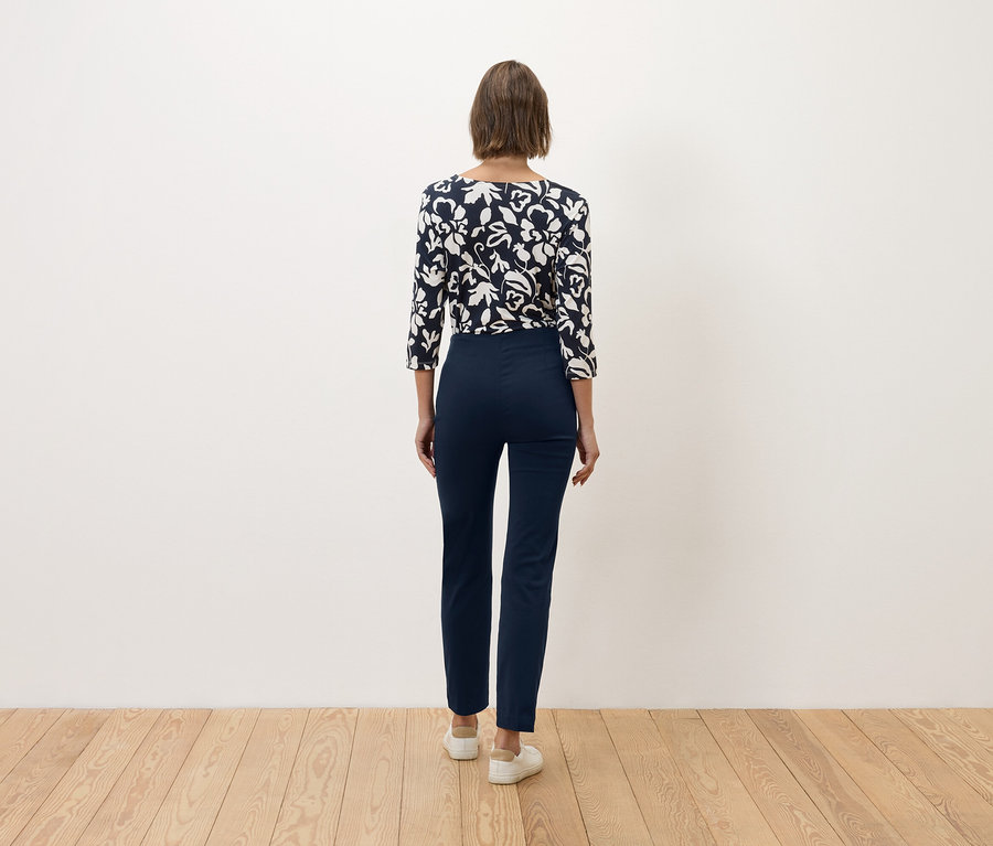 Žena s hnědými vlasy stojí zády ke kameře a má na sobě tmavě modré kalhoty a modro-bílý květinový top.
