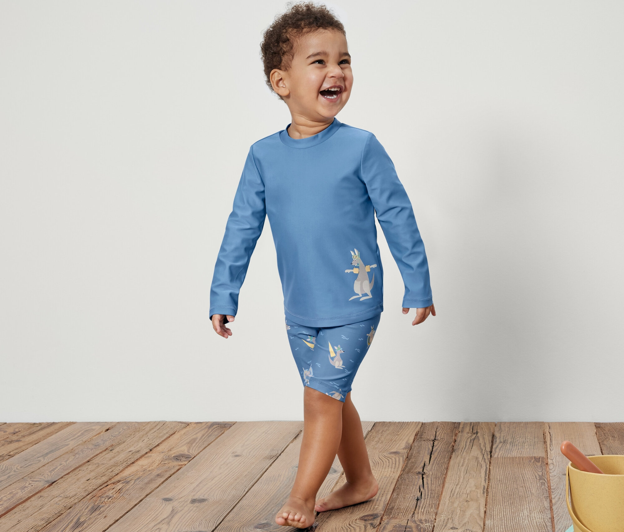 Usmívající se chlapeček v modré dětské soupravě s UV faktorem 80 s potiskem klokana kráčí po dřevěné podlaze. Vpravo je žlutý kyblík s hračkami na písku.