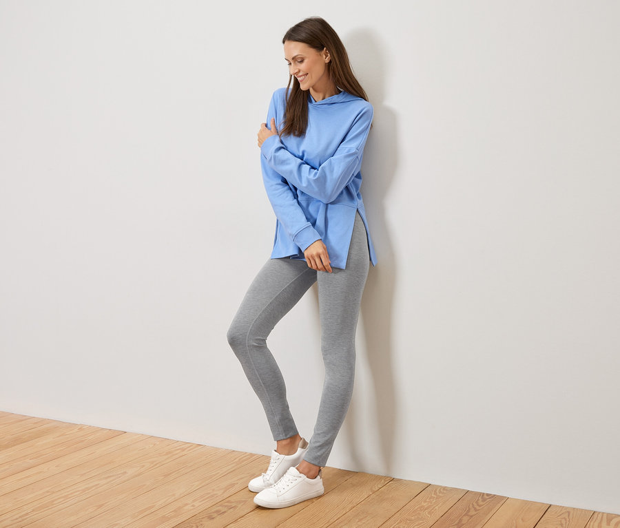 Žena se opírá o zeď, má na sobě světle modrou mikinu s kapucí s bočními rozparky, šedé legíny a bílé tenisky.