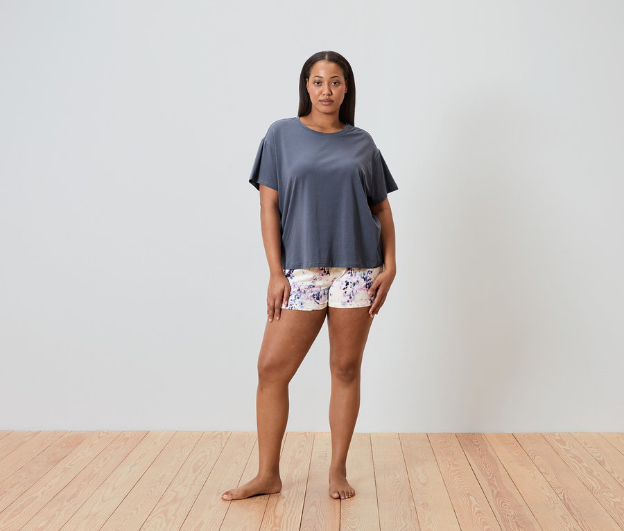Žena stojí bosá na dřevěné podlaze a má na sobě šedé tričko a šortky s květinovým vzorem.