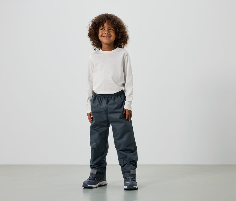 Usmívající se chlapec s kudrnatými vlasy má na sobě tričko s dlouhým rukávem, nepromokavé kalhoty a tenisky.