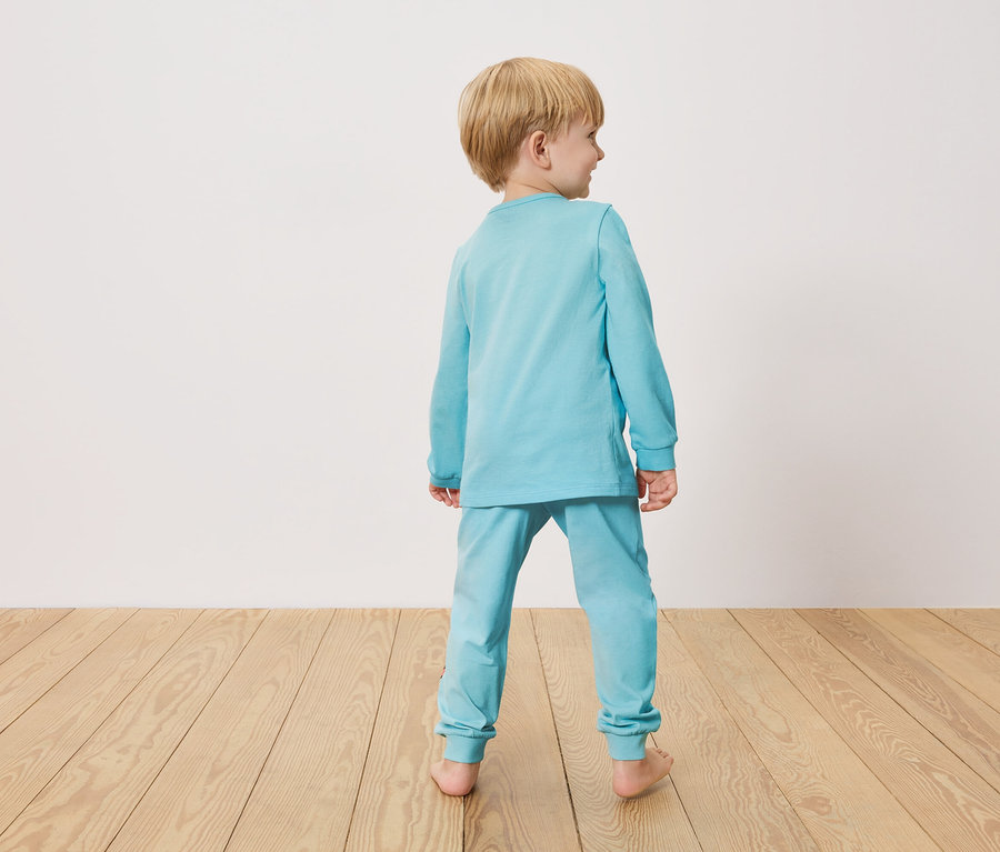Malý chlapec zezadu, oblečený ve světle modrém pyžamu, stojí na dřevěné podlaze před bílou stěnou.