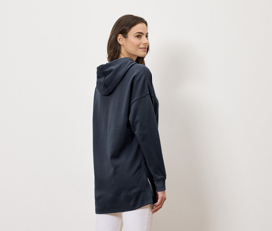 Žena nosí dlouhou tmavě modrou mikinu s kapucí a bílé kalhoty, boční pohled.