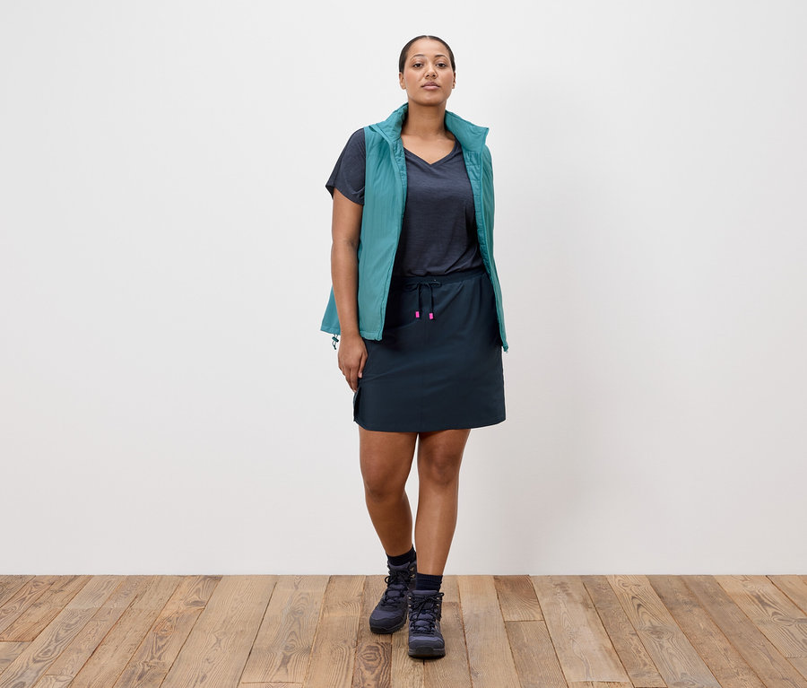 Žena stojí před bílou stěnou, má na sobě tmavě modrou sukni, tmavě modré tričko, petrolejovou vestu a tmavě modré trekové boty.