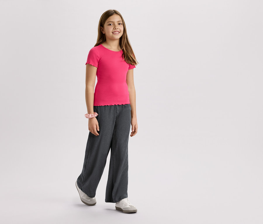 Dívka má na sobě růžové tričko, šedé kalhoty a tenisky na bílém pozadí.