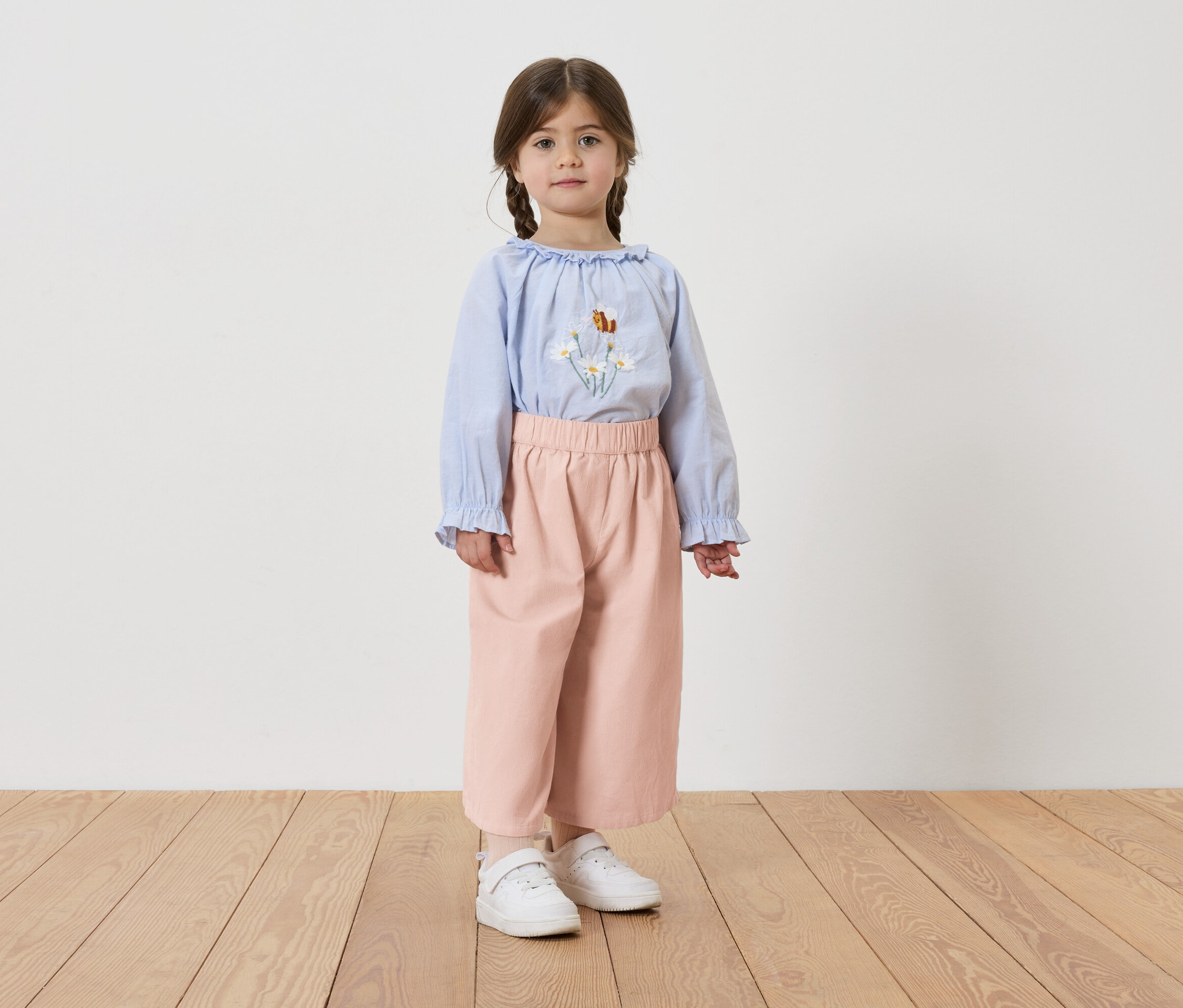 Dívka s copy nosí světle modrou halenku s květinovou výšivkou, růžové kalhoty a bílé tenisky.